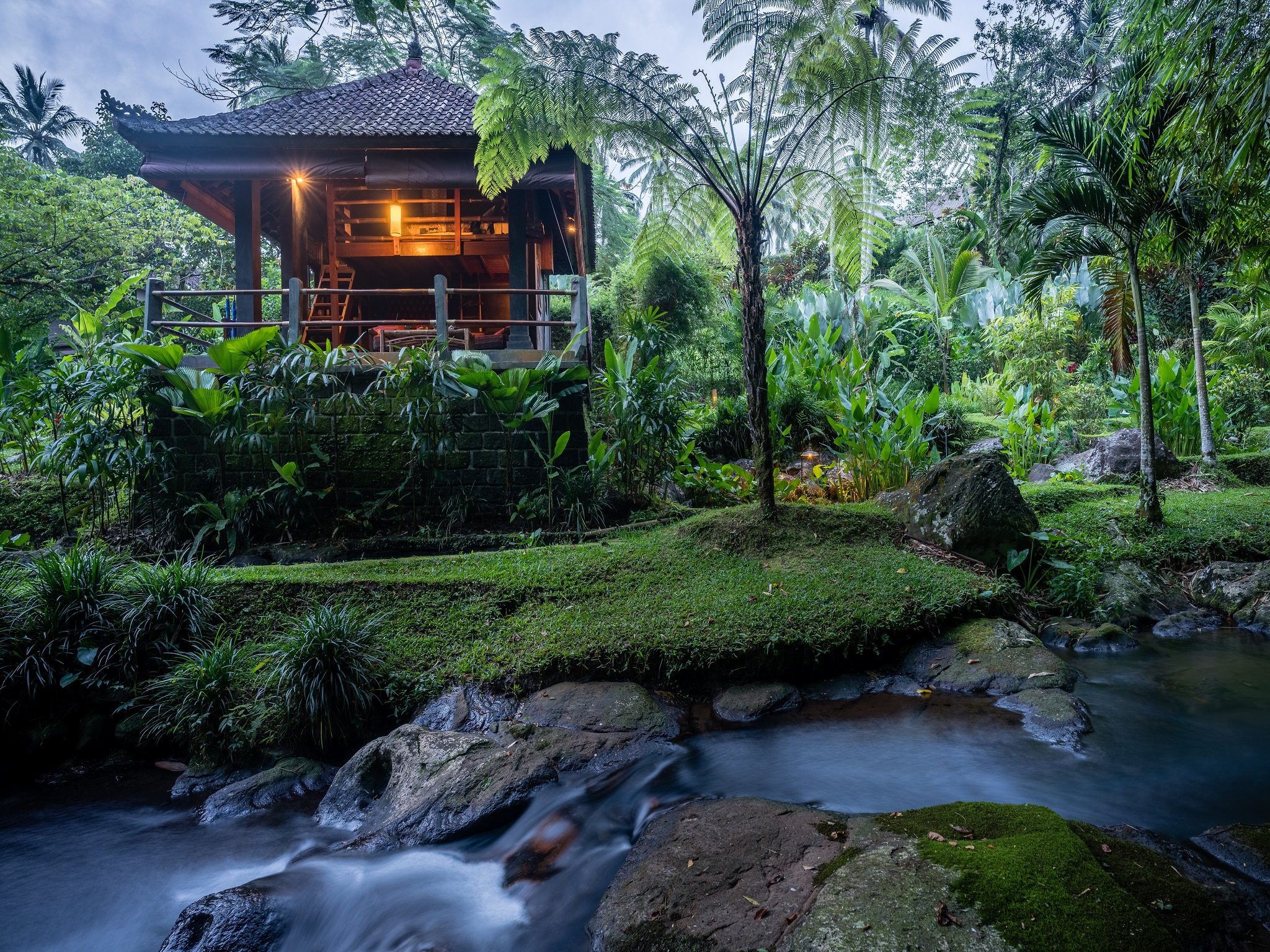 The Buddha Bungalow lit at evening, its open verandah looking over a rocky mountain stream, with tree ferns and palms crowding the banks.